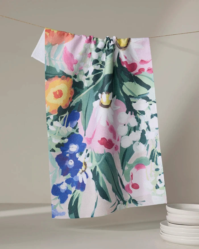 The Ania Zwara Art "Garden Bouquet" tea towel features vibrant pink, yellow, blue, and green blooms on a white ultra-absorbent recycled fabric, hanging above a stack of plates for a colorful kitchen accent.