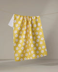 The Sara Mari Garden Crush kitchen towel, featuring a white and pink flower pattern on yellow eco-friendly fabric, hangs on a clothesline against a plain background.