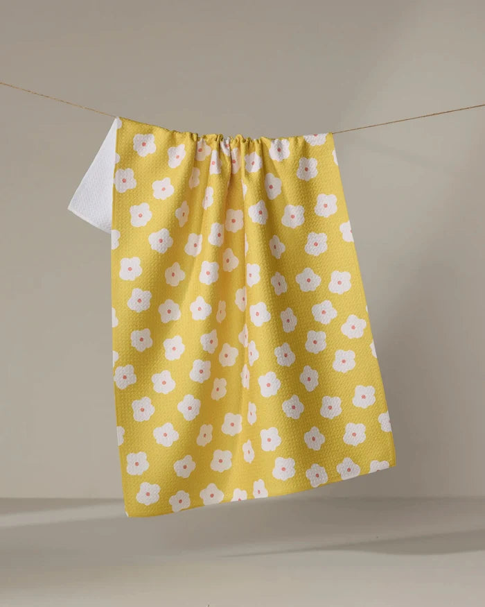 The Sara Mari Garden Crush kitchen towel, featuring a white and pink flower pattern on yellow eco-friendly fabric, hangs on a clothesline against a plain background.