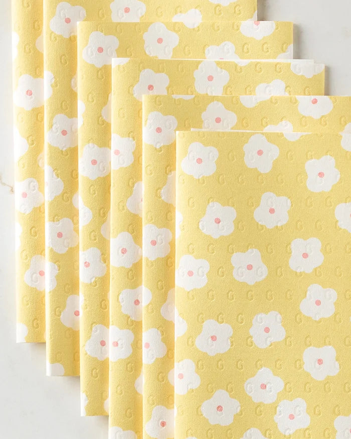 Five yellow Sara Mari "Garden Crush" eco-friendly kitchen towels featuring a repeating white flower pattern with pink centers are fanned out on a light surface.