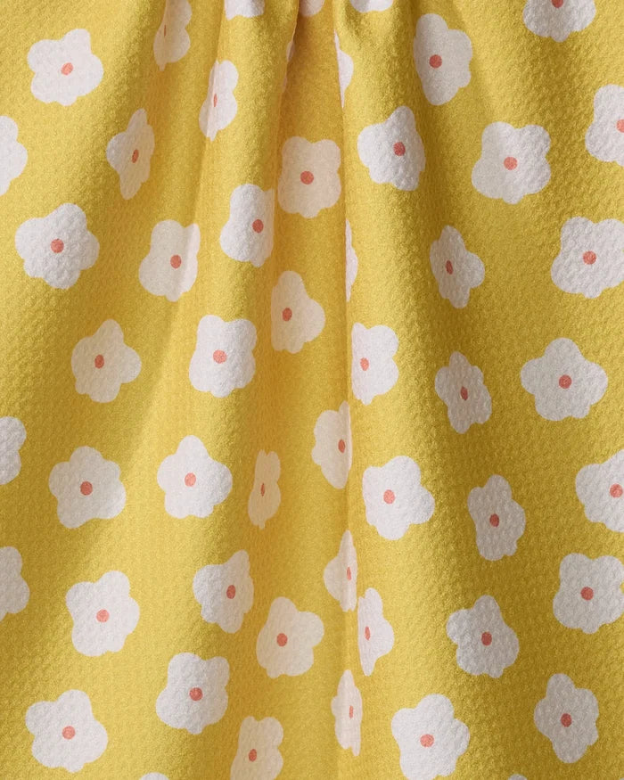 The Sara Mari "Garden Crush" kitchen towel features yellow textured fabric with a repeating white four-petaled flower pattern, each centered by a pink dot. Eco-friendly and stylish, it drapes softly, creating gentle folds.