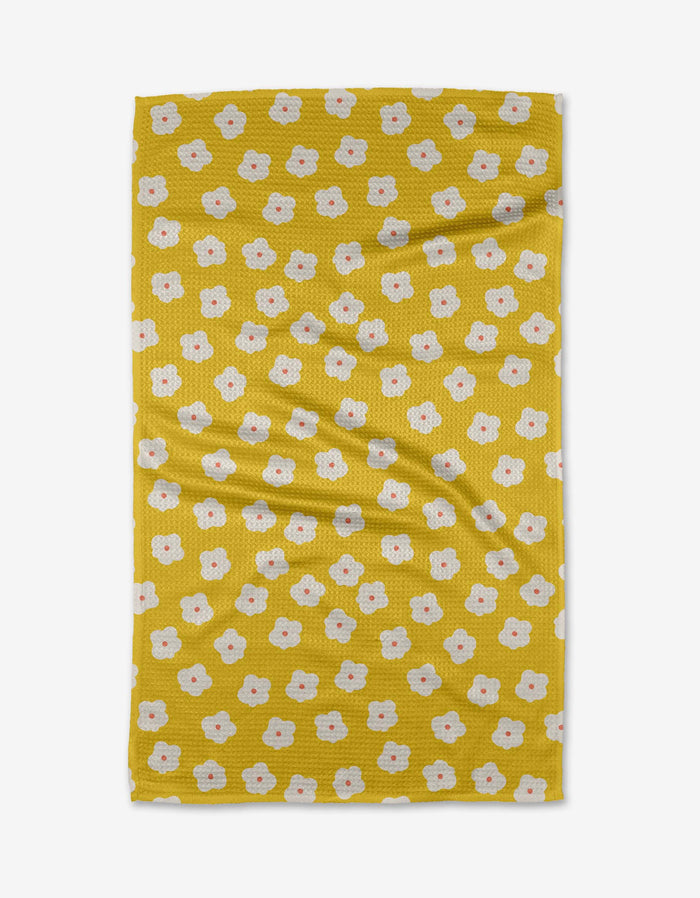 The Sara Mari “Garden Crush” tea towel features a yellow base with scattered small white flowers with orange centers. Laid flat, its gentle folds highlight its eco-friendly recycled material construction.