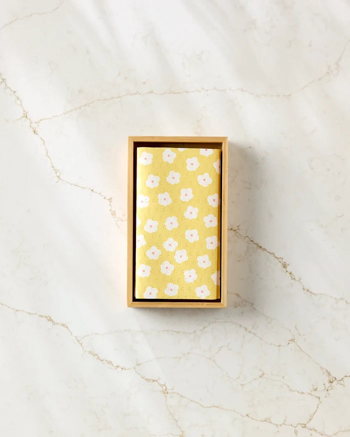 A rectangular Sara Mari wooden box holds the Garden Crush yellow eco-friendly kitchen towel with a white and pink floral pattern, displayed on a light marble surface with subtle veining.