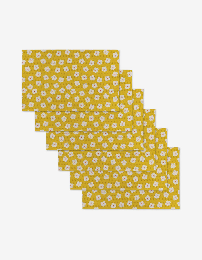 Six Sara Mari Garden Crush placemats, rectangular and yellow with a floral pattern of white flowers and green leaves, made from eco-friendly materials, are stacked in a slightly overlapping arrangement on a white background.