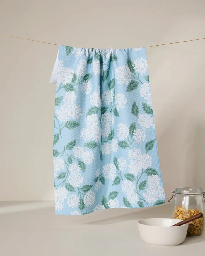 The GEOMETRY Garden Of Edith blue tea towel, featuring a white floral and green leaf pattern, hangs on a string above a white bowl and pasta-filled jar. Made from quick-drying, recycled materials.