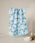 The GEOMETRY Garden Of Edith blue tea towel, featuring a white floral and green leaf pattern, hangs on a string above a white bowl and pasta-filled jar. Made from quick-drying, recycled materials.