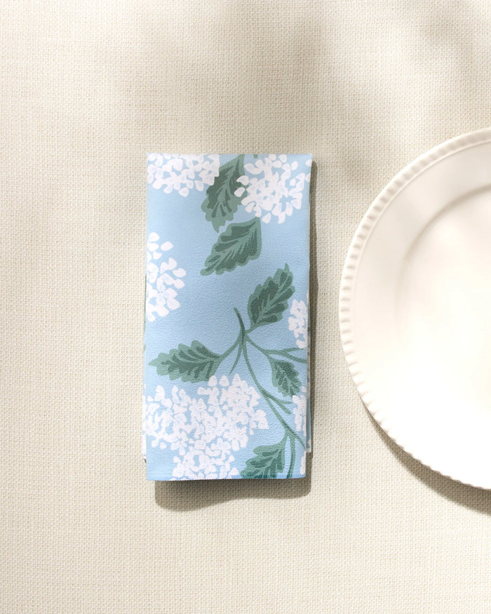 The GEOMETRY 'Garden Of Edith' reusable napkin, featuring a blue background with white florals and green leaves, is shown folded on a beige textured surface beside a white scalloped-edge plate—an elegant dinner napkin set.
