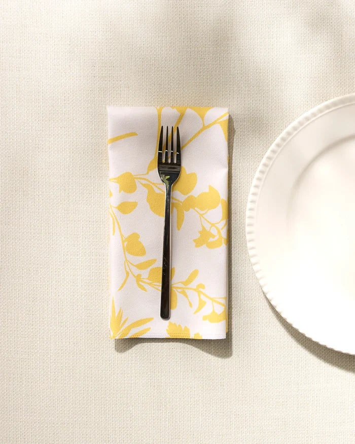 A silver fork rests on GEOMETRY's "Garden of Gold" yellow and white floral washable napkin, folded beside a white plate on a textured, light-colored tablecloth.