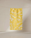 The GEOMETRY Garden of Gold bar towel, made from recycled materials, features a white botanical leaf and herb pattern on yellow and hangs on a string against a plain light background.