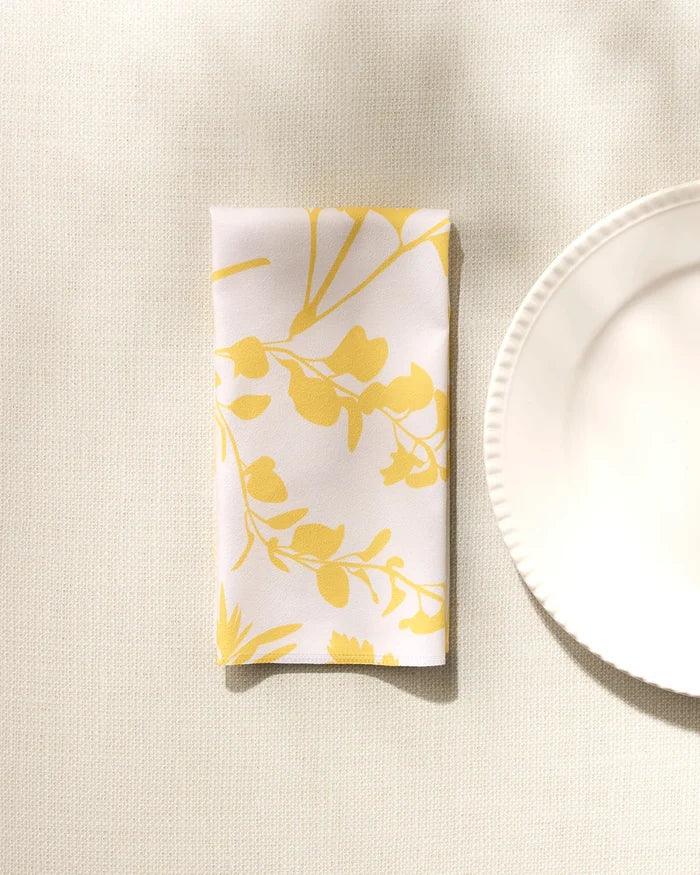 The GEOMETRY "Garden of Gold" folded white washable cloth napkin, featuring a yellow floral pattern, rests beside a scalloped-edge white plate on a light textured surface under soft natural light.