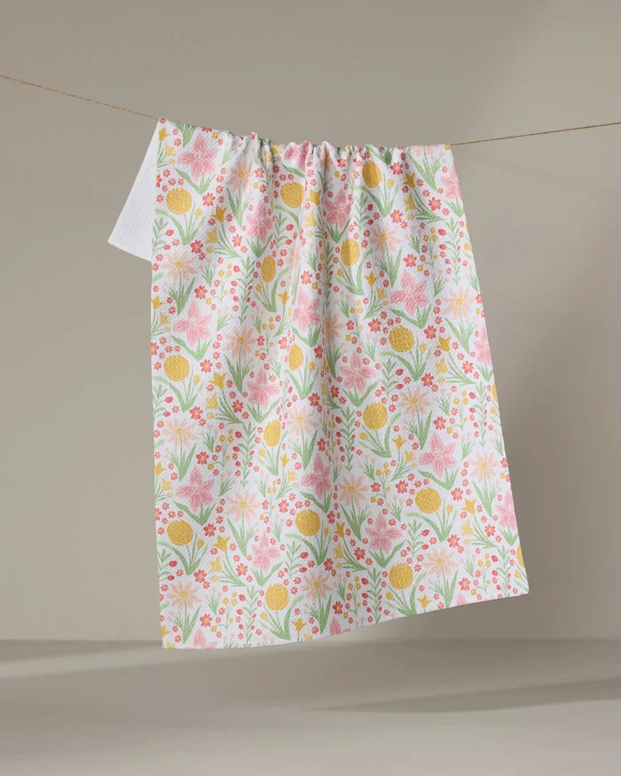 The “Garden of Light” tea towel by Danelys Sidron, featuring a quick-drying fabric and colorful pink, yellow, and green floral patterns, hangs on a clothesline against a neutral background.