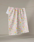 The “Garden of Light” tea towel by Danelys Sidron, featuring a quick-drying fabric and colorful pink, yellow, and green floral patterns, hangs on a clothesline against a neutral background.