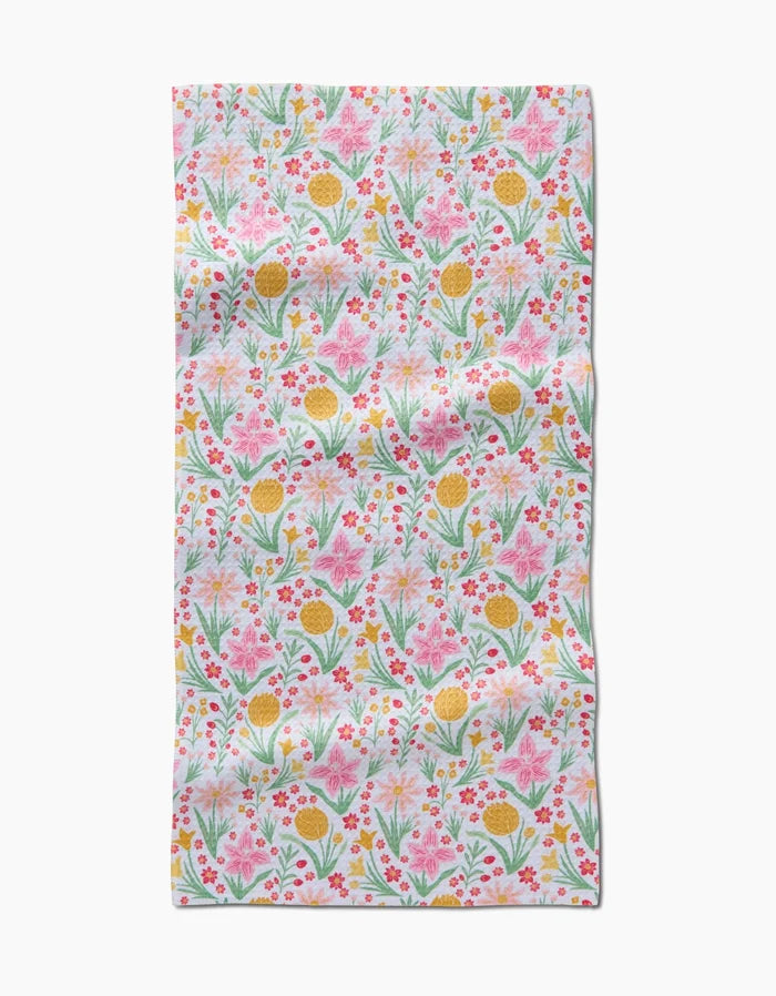 The Garden of Light bar towel by Danelys Sidron features a colorful floral pattern—pink, yellow, and orange flowers with green leaves—on white Geoweave fabric made from recycled materials. The material has a slightly wrinkled look.