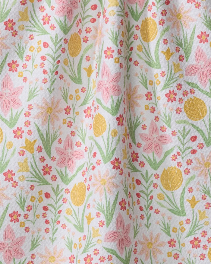 A close-up of the "Garden of Light" tea towel by Danelys Sidron, showcasing colorful yellow, pink, and orange flowers with green leaves on white fabric. Soft folds highlight its quick-drying quality.