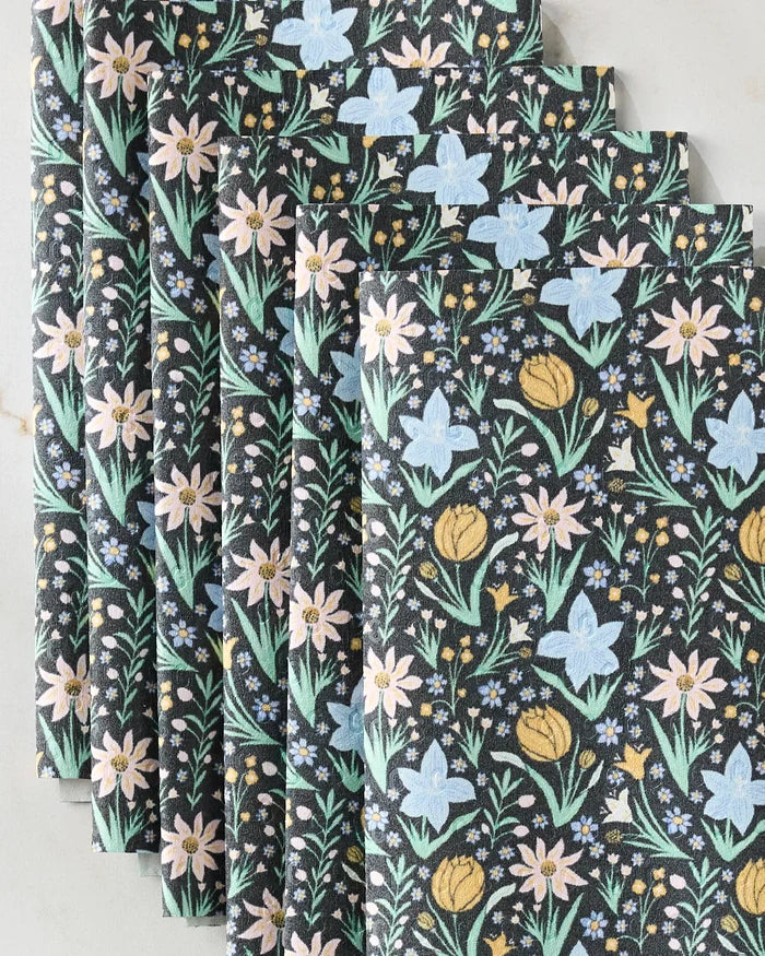 A stack of Danelys Sidron's Garden of Light reusable paper towels features a floral pattern with yellow tulips, blue and pink flowers, and green leaves on a dark background, arranged diagonally on a light surface.