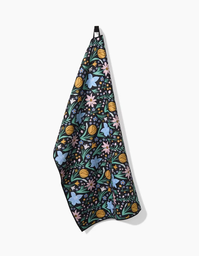 The Danelys Sidron Garden of Light hanging tea towel features a black background with vibrant blue, yellow, pink, and green floral patterns, and is thoughtfully crafted from post-consumer recycled materials.