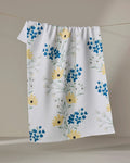 The Ashley Celena "Garden Picks" tea towel, in white with a textured pattern, hangs on a line and features a floral design of yellow and blue flowers with green stems.
