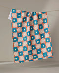 The Garden Plot tea towel by Kelsey Weigl features a retro floral pattern in blue, white, and peach. Made from recycled materials, it’s an eco-friendly choice for your kitchen and is shown hanging against a light background.