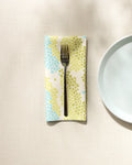 A Geo Shell napkin by GEOMETRY with blue and green dots rests under a fork on a cream tablecloth, beside a plain white plate.
