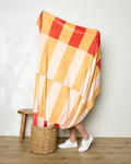 A person stands hidden behind the Geometry Geometric Sunset throw blanket in red, yellow, and peach tones. Only their lower legs and white sneakers appear next to a wooden stool and woven basket on a wooden floor.