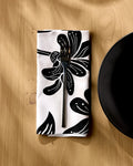 A fork rests on a neatly folded Geometry Giant Lush Floral reusable napkin, featuring an eco-friendly black and white print, atop a light wooden table with a black plate's edge visible to the right.
