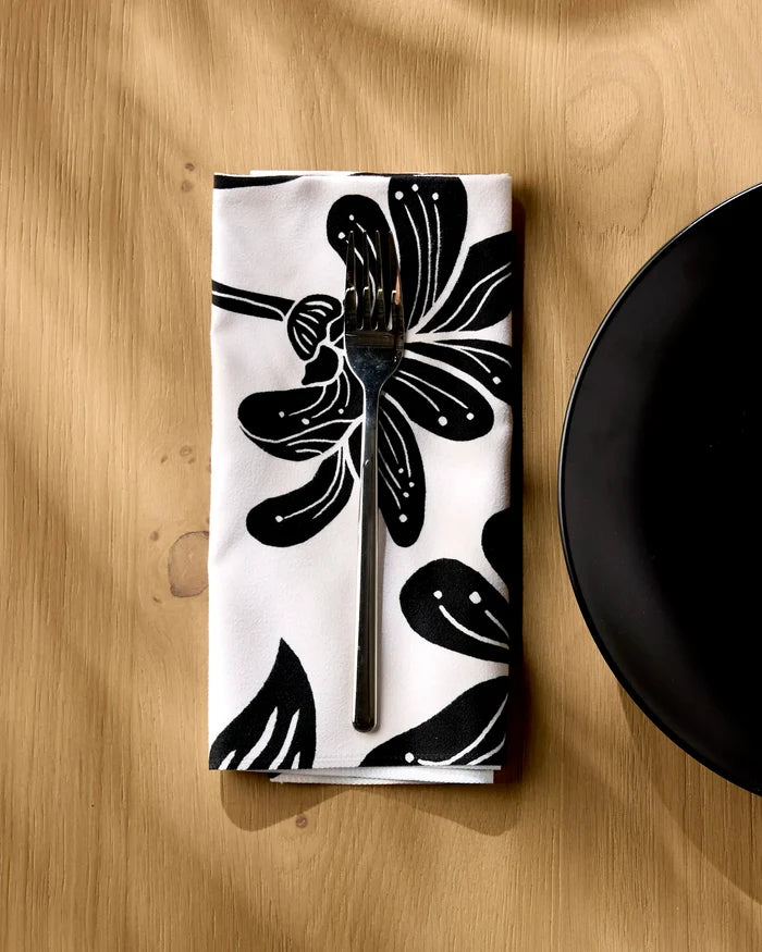 A fork rests on a neatly folded Geometry Giant Lush Floral reusable napkin, featuring an eco-friendly black and white print, atop a light wooden table with a black plate's edge visible to the right.