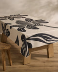 A rectangular wooden table is draped with Geometry’s Giant Lush Floral cream tablecloth, showcasing bold black abstract florals. A rustic stool sits beside the table on a light wood floor.