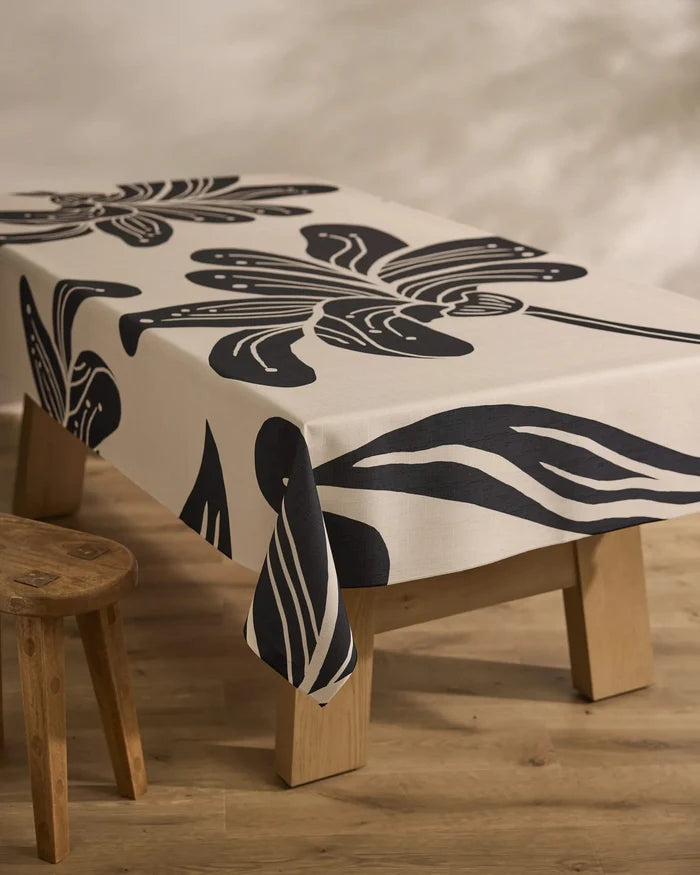 A rectangular wooden table is draped with Geometry’s Giant Lush Floral cream tablecloth, showcasing bold black abstract florals. A rustic stool sits beside the table on a light wood floor.