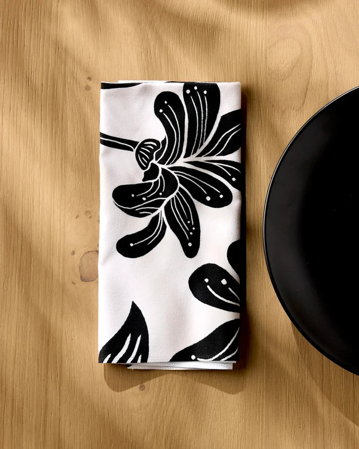The Geometry Giant Lush Floral folded white reusable dinner napkin, featuring a bold black floral pattern, rests on a light wooden table beside the edge of a round black plate.