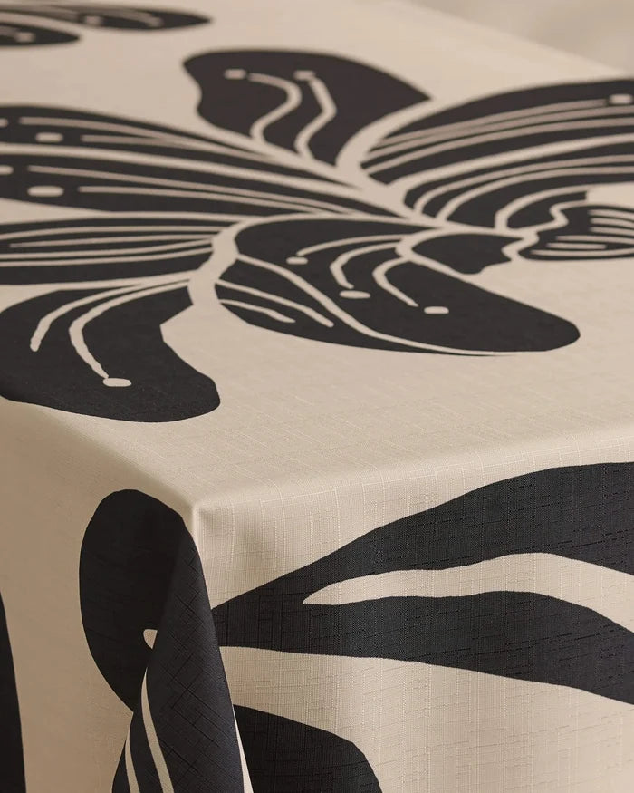 A close-up of a table featuring Geometrys Giant Lush Floral tablecloth, cream-colored and stain-resistant, decorated with a bold abstract black leaf design that extends over the edge.