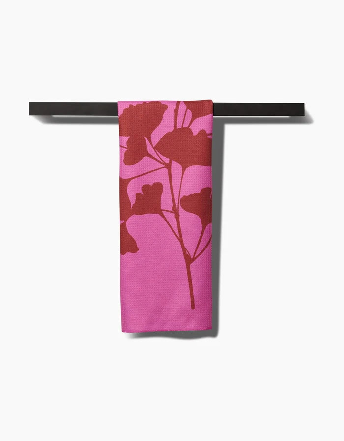 A PrintedPlant Ginkgo towel, featuring a bold red botanical print on pink ultra-absorbent Geoweave fabric, hangs neatly from a dark bar against a white background.
