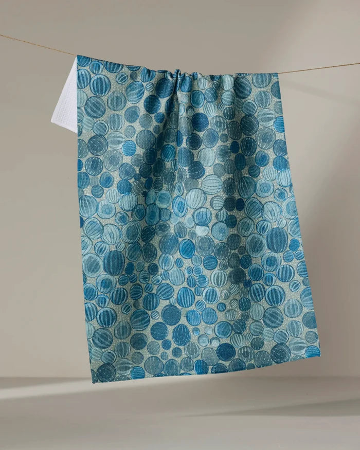 A blue ultra-absorbent tea towel, Glass Baubles by Katie O'Shea, featuring variously shaded blue circles, hangs on a clothesline against a neutral background.