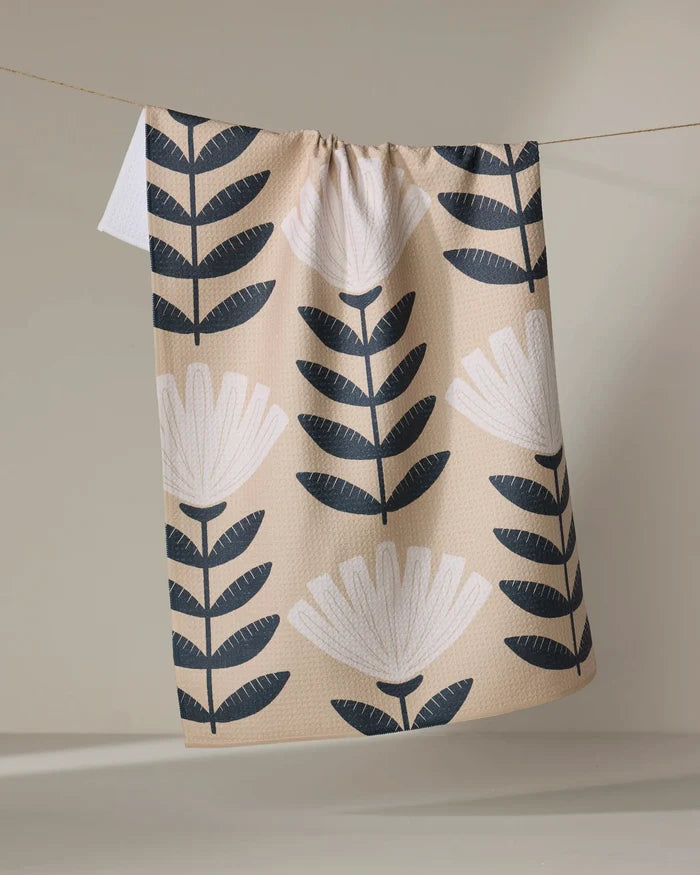 The Gloria kitchen tea towel by Amy MacCready, crafted from recycled materials, displays large black and white stylized flowers and leaves on beige fabric, hanging on a clothesline against a neutral backdrop.