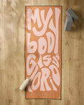 The peach-colored "Glorious" yoga mat by Arielle Estoria, featuring bold wavy pink MY BODY IS GLORIOUS text, is displayed on a light wood floor with a white water bottle, gray yoga blocks, and a non-slip towel for comfort and stability.