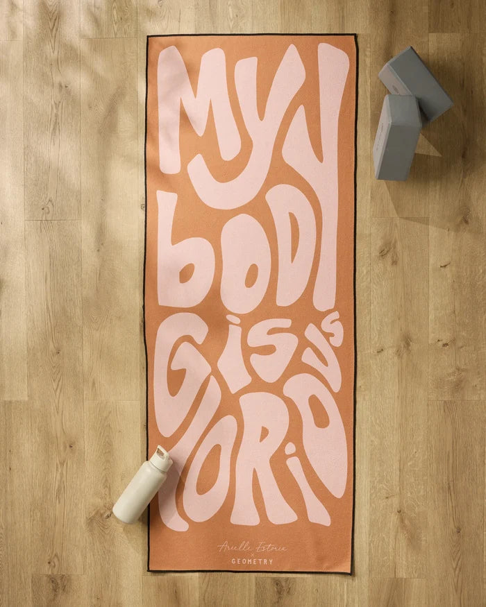 The peach-colored "Glorious" yoga mat by Arielle Estoria, featuring bold wavy pink MY BODY IS GLORIOUS text, is displayed on a light wood floor with a white water bottle, gray yoga blocks, and a non-slip towel for comfort and stability.