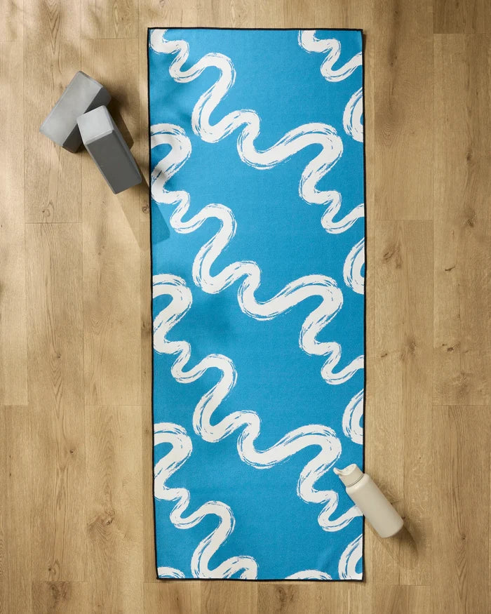 The "Go With the Flow" yoga mat by Elena Fay, featuring blue with white wavy lines, is set on a wooden floor alongside a non-slip towel, two gray blocks, and a white water bottle.