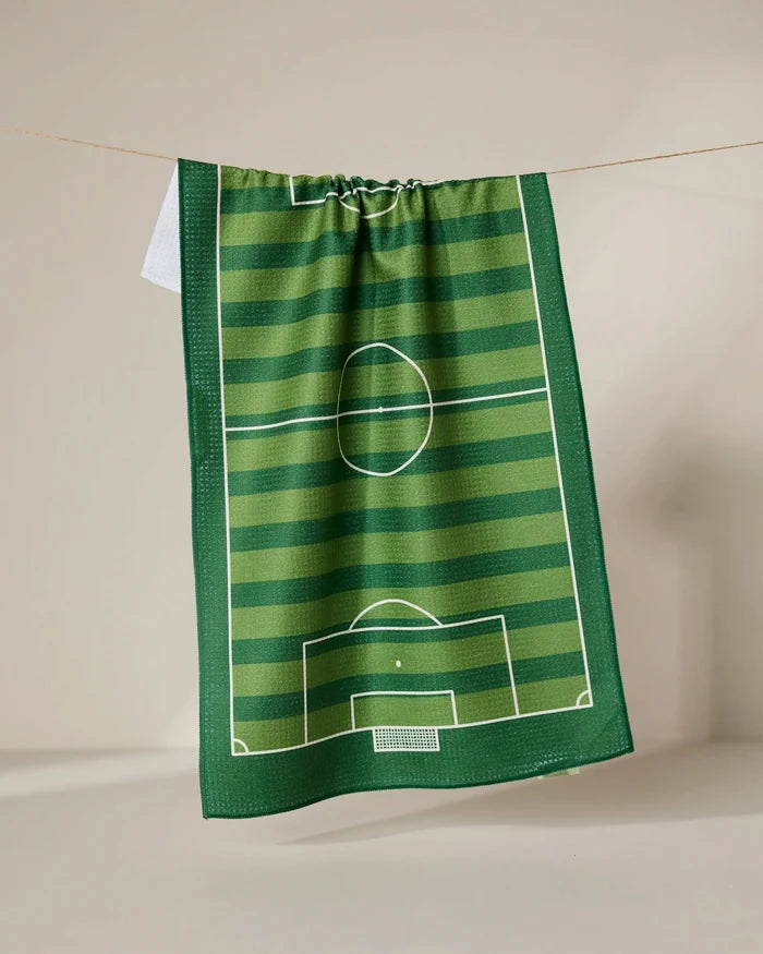The GEOMETRY Goal tea towel features a green soccer field design with white markings and stripes, hanging from a string against a neutral backdrop.