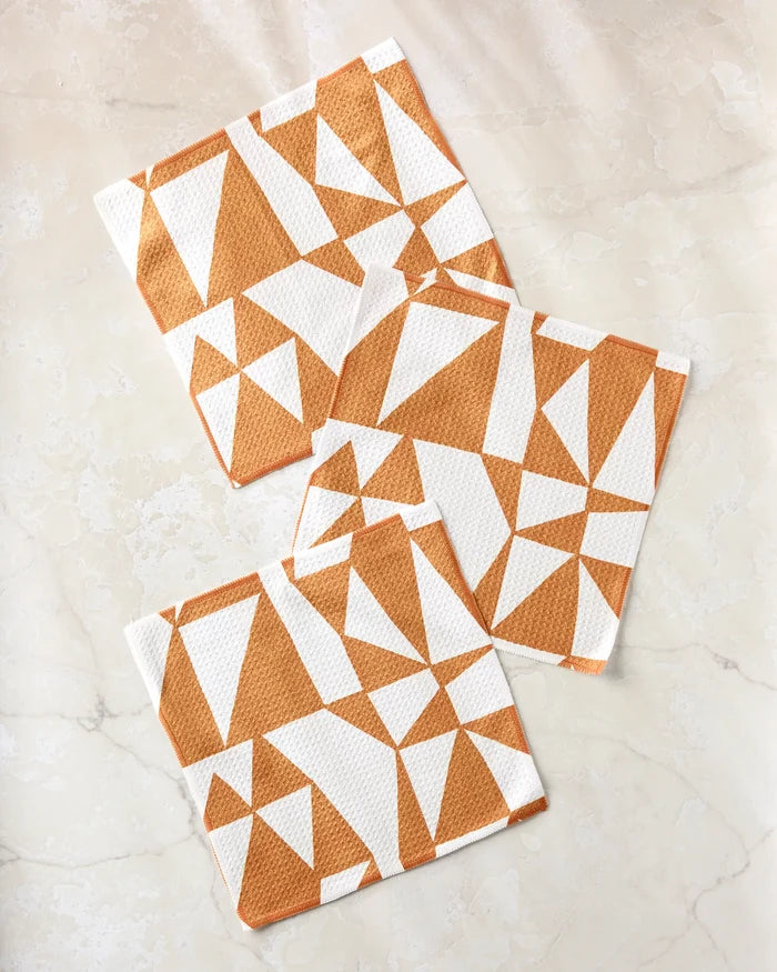 Three white super-absorbent Lisa Nelson dishcloths from the Golden Angles collection, featuring orange geometric triangle patterns, are arranged on a light beige marble surface.