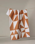 The "Golden Angles" tea towel by Lisa Nelson, featuring a geometric white and terracotta triangle pattern and crafted from recycled materials, hangs on a clothesline against a neutral background.