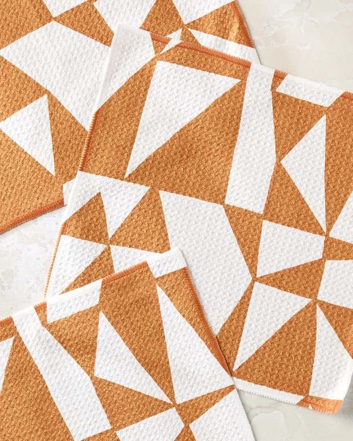 Four "Golden Angles" cloth napkins by Lisa Nelson, featuring an orange and white abstract geometric triangle pattern, overlap on a light surface—an eco-friendly alternative to standard dishcloth sets.