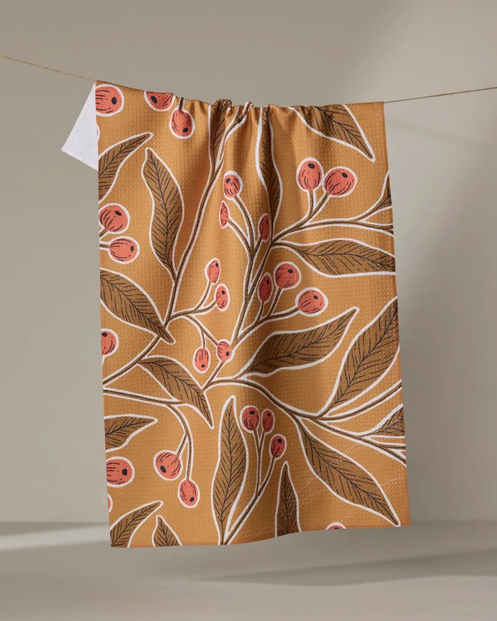 The "Golden Berries" tea towel by Amy MacCready, made from recycled materials, features an illustrated design of leafy branches with red berries and white outlines on a brown background.
