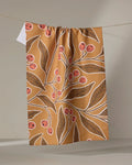 The "Golden Berries" tea towel by Amy MacCready, made from recycled materials, features an illustrated design of leafy branches with red berries and white outlines on a brown background.