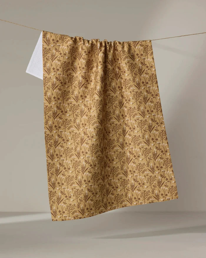 The "Golden Fall Flowers" tea towel by Danelys Sidron, featuring a brown floral pattern on beige recycled fabric, hangs from a clothesline against a neutral background, with part of a small white towel visible behind it.