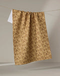 The "Golden Fall Flowers" tea towel by Danelys Sidron, featuring a brown floral pattern on beige recycled fabric, hangs from a clothesline against a neutral background, with part of a small white towel visible behind it.