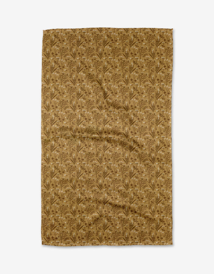 The Golden Fall Flowers tea towel by Danelys Sidron is a rectangular, brown cloth made from recycled materials, featuring a repeating botanical pattern of small leaves and branches. It is slightly wrinkled and shown on a white background.