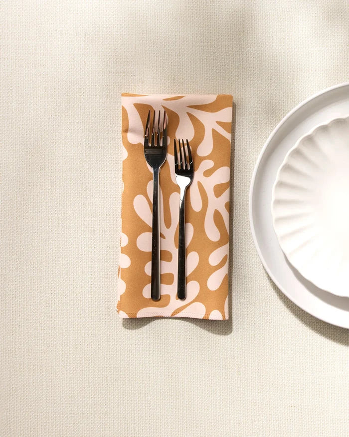 Two forks rest side by side on a folded GEOMETRY Golden Fall reusable dinner napkin with a beige and white abstract pattern, next to a scalloped white plate on a light textured surface.
