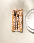 Two forks rest side by side on a folded GEOMETRY Golden Fall reusable dinner napkin with a beige and white abstract pattern, next to a scalloped white plate on a light textured surface.