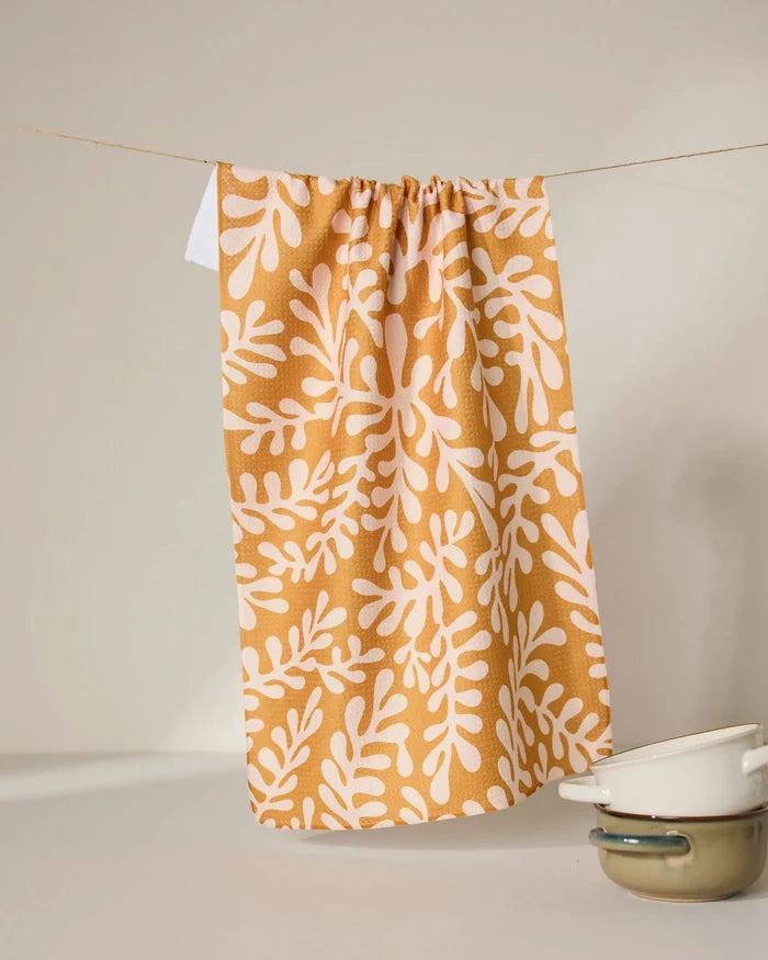 The GEOMETRY Golden Fall tea towel, crafted from recycled materials in mustard yellow with a white leafy pattern, hangs on a string. In front, two cream enamel pots with dark rims are stacked against a neutral background.