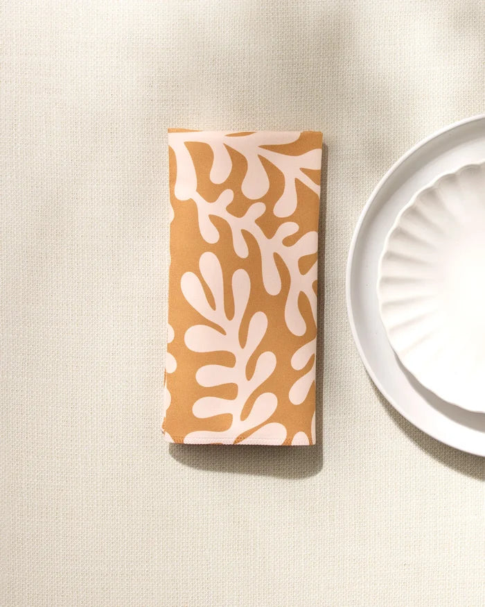 The GEOMETRY Golden Fall recycled fabric napkin, featuring a beige and white abstract leafy pattern, is folded beside a white scalloped-edge plate on a lightly textured tablecloth.