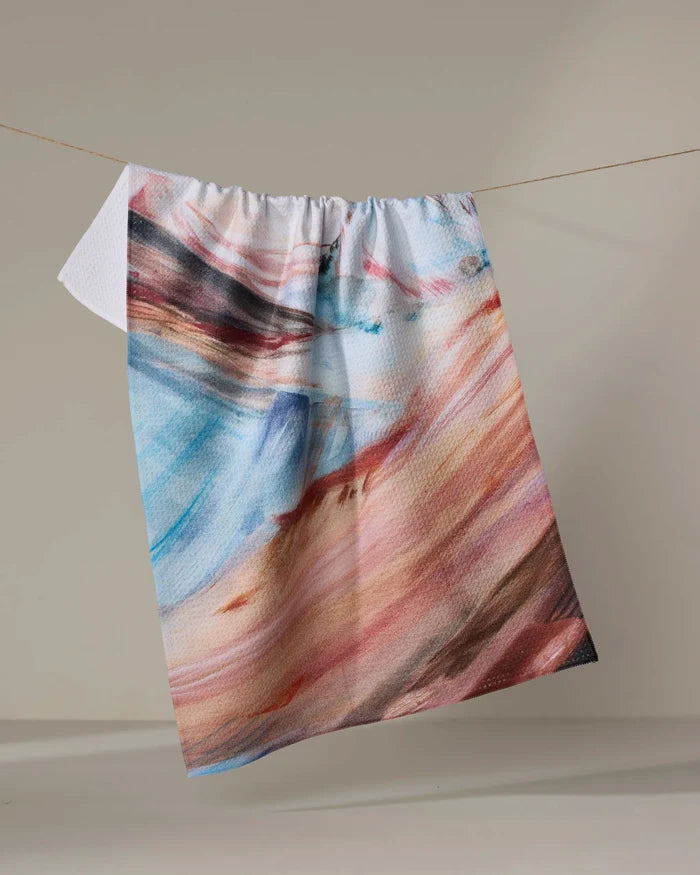 The "Golden Hour in the Rockies" tea towel by Kelly Peters features an abstract, colorful pattern in blue, pink, orange, and brown. Made from recycled materials for a sustainable touch, it hangs on a clothesline against a plain light background.
